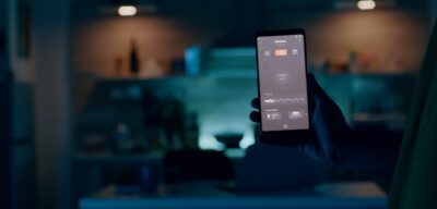 Person holding mobile phone with high tech application in smart house features controlling lights with wireless device. Man turning on the light sitting in home kitchen with automation lighting system